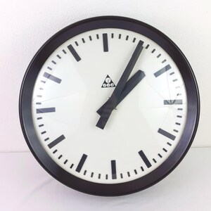 Puede incluir: Un reloj de pared redondo con esfera blanca y manecillas y marcadores negros. El reloj tiene un marco marrón oscuro y un pequeño logotipo en el centro. Las manecillas del reloj señalan aproximadamente las 2:10.
