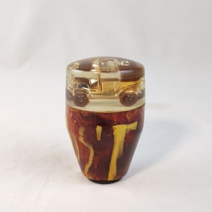 May include: A cylindrical gear shift knob featuring a clear resin top section that encapsulates a small gold-colored vintage car model. The lower portion of the knob is crafted from a marbled resin material with swirling patterns of deep brown and yellow.
