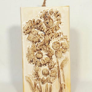 Può includere: Un'applique da parete rettangolare in ceramica con un motivo floreale in rilievo nei toni del marrone e del beige. Un anello in fibra naturale è attaccato nella parte superiore per appenderlo. L'opera presenta un motivo botanico strutturato e tridimensionale.