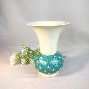 Peut inclure: Un vase décoratif avec un dessus crème et une base turquoise ornée de motifs floraux jaunes. Le vase a une bordure dorée et une ouverture évasée. Une branche de fleurs blanches repose à côté.