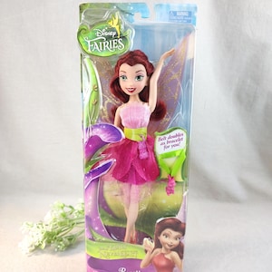 May include: A Disney Fairies Rosetta doll in packaging. The doll has red hair, a pink top, and a layered pink skirt. The packaging includes the Disney Fairies logo and the text "Belt doubles as bracelet for you!"