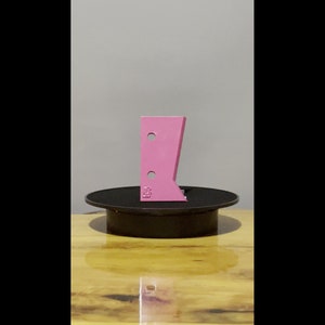May include: A pink plastic 3D printed object with two holes on a black rotating platform. The object is shaped like a wedge with a rounded top.