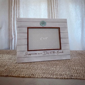 May include: White wooden picture frame with a seashell design. The frame is 4 inches by 6 inches and has the text "Happiness is a Day at the Beach" on the bottom.