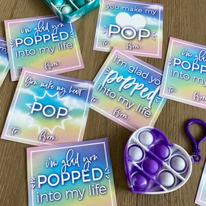 May include: A set of colorful Valentine's Day cards with the text "I'm glad you popped into my life" and "You make my heart pop". The cards feature a rainbow tie-dye background and a heart shape with a pop-it toy.