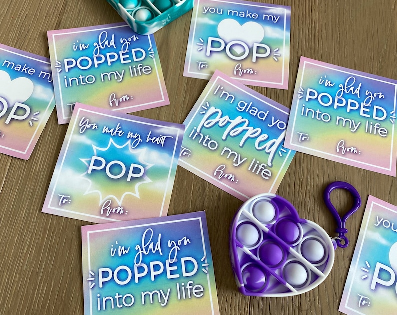 May include: A set of colorful Valentine's Day cards with the text "I'm glad you popped into my life" and "You make my heart pop". The cards feature a rainbow tie-dye background and a heart shape with a pop-it toy.