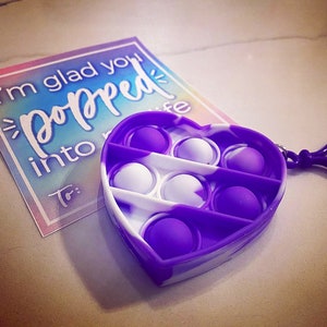 May include: Purple and white heart-shaped pop it fidget toy with a keychain. The toy has 7 dimples that can be pushed in and out. The text on the card reads "I'm glad you popped into my life."