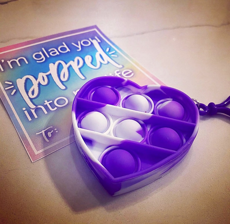 May include: Purple and white heart-shaped pop it fidget toy with a keychain. The toy has 7 dimples that can be pushed in and out. The text on the card reads "I'm glad you popped into my life."