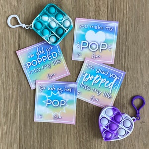 May include: Four colorful Valentine's Day cards with the word "POP" and the phrase "I'm glad you popped into my life". The cards have a rainbow gradient background. Two of the cards have a heart shape with the word "POP" inside. The cards are next to a blue and white pop-it toy and a purple and white heart-shaped pop-it toy.
