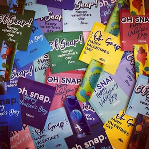May include: A collection of colorful Valentine's Day gift tags with the phrase "Oh Snap!" and "Happy Valentine's Day". The tags are printed on various colored cardstock and feature different designs, including a dinosaur, a rocket ship, and a heart.