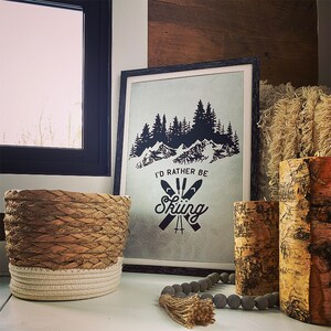 May include: A black framed print with a white background and a black silhouette of a mountain range with trees. The text "I'd rather be skiing" is printed in black with a graphic of two crossed skis. The print is sitting on a white surface with a brown and white woven basket, a gray beaded garland, and two birch bark cylinders.