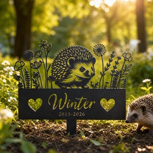May include: Black metal garden stake with a cut-out hedgehog design and floral details. The stake displays the name "Winter" and the years 2015-2026, with heart-shaped cutouts. A real hedgehog is in the foreground.