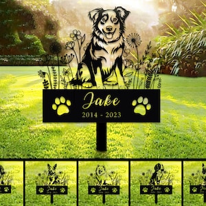 Op de afbeelding: Zwarte metalen silhouet van een hond met bloemen en de tekst "Jake 2014 - 2023" op een zwart bord met poot afdrukken. Het bord is bevestigd aan een metalen paal om in de grond te steken.