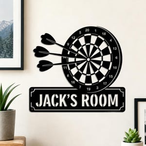 May include: A black metal wall sign featuring a dartboard graphic with three darts embedded in the centre. Below the dartboard is a rectangular plaque with rounded corners and the white text "JACK'S ROOM" in a bold, sans-serif font.