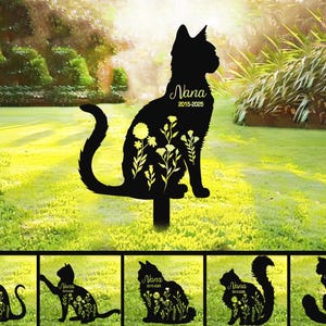 Op de afbeelding: Zes zwarte metalen silhouetten van katten met bloemen en de naam "Nana" en de data "2015-2023" op een van de silhouetten. De silhouetten bevinden zich op een groene grasachtige achtergrond.
