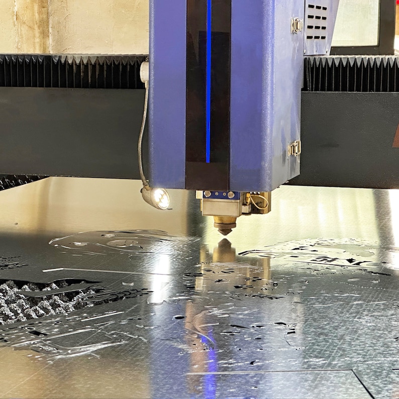 May include: A close-up of a laser cutter cutting a metal sheet. The laser cutter is blue and has a light on the side. The metal sheet is silver and has a design cut into it.