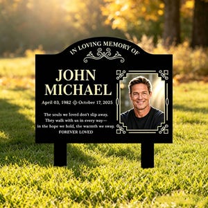 Puede incluir: Una placa conmemorativa rectangular negra para jardín con texto dorado IN LOVING MEMORY OF JOHN MICHAEL, las fechas 03 de abril de 1982 al 17 de octubre de 2025 y un poema. Incluye un retrato enmarcado de una persona sonriente y dos estacas negras.