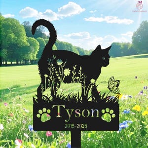 Op de afbeelding: Zwarte metalen katten silhouet tuinpaal met de naam "Tyson" en de jaren 2015-2025. De kat loopt door gras en bloemen, met een vlinder in de buurt. De achtergrond is een groen veld en een blauwe lucht.