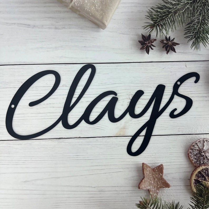 May include: Black metal sign with the word "Clays" written in a cursive font. The sign is on a white wooden background.