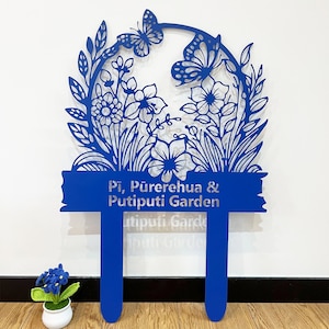 Op de afbeelding: Blauwe metalen tuinmarker met een bloemmotief en de tekst "Pi, Purehua & Putiputi Garden".