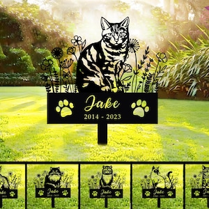 Op de afbeelding: Een zwarte metalen tuinpaal met een katten silhouet, bloemen en de naam "Jake" met de jaren 2014-2023. De paal bevat pootafdrukken. Extra palen met verschillende kattenrassen worden hieronder getoond.