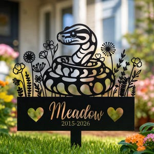 Può includere: Paletto da giardino in metallo nero con un design stilizzato di serpente e dettagli floreali. Il paletto include il nome "Meadow" e gli anni 2015-2026, con ritagli a forma di cuore. Il design è ambientato su uno sfondo di erba verde e fiori colorati.