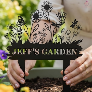 Könnte beinhalten: Schwarzer Metall-Gartenstecker mit floralem Design, darunter Sonnenblumen, Schmetterlinge und Libellen. Der Text "JEFF'S GARDEN" ist unten ausgeschnitten. Der Stecker wird in Erde gehalten.