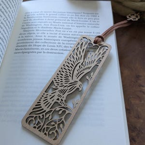 Peut inclure: Un marque-page en bois avec un motif découpé au laser d'un oiseau perché sur une branche. Le marque-page est brun clair et présente un design délicat et complexe.