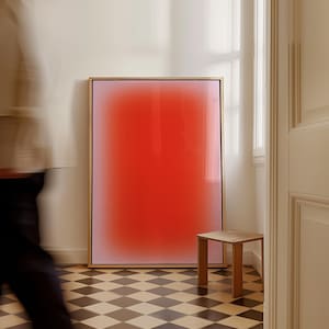 May include: A framed abstract art print with a gradient of pink to red. The print is hanging on a white wall in a room with a checkered floor.