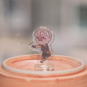 May include: A clear acrylic figurine of a cartoon character in a yellow and black uniform, with bandages on their head. The character has a distressed expression. The figurine is on a clear base, set on a light orange surface.