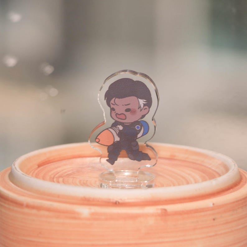 May include: A clear acrylic figurine of a cartoon character in a running pose, holding an orange and blue object. The character has dark hair with a white streak and is wearing a dark blue outfit. The figurine is on a clear base, set on a light orange, circular surface.