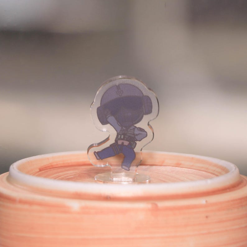 May include: A clear acrylic figurine of a cartoon character in a blue and black uniform, posed in a dynamic stance. The figurine is mounted on a clear base and set against a blurred background. The figurine is placed on a peach-colored ceramic container.