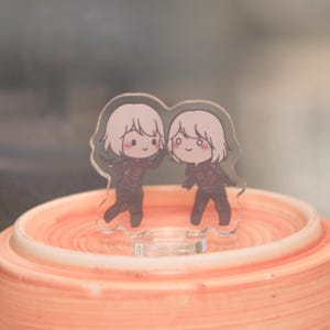 May include: A clear acrylic charm featuring two cartoon figures with white hair and dark outfits. The figures are smiling and waving. The charm is standing on a peach-colored surface.