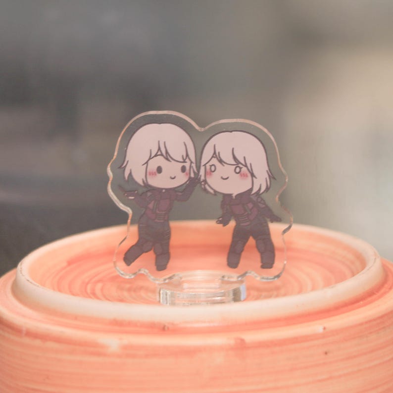 May include: A clear acrylic charm featuring two cartoon figures with white hair and dark outfits. The figures are smiling and waving. The charm is standing on a peach-colored surface.