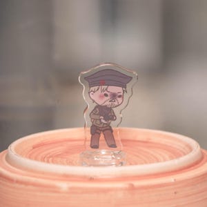May include: A clear acrylic figurine of a cartoon soldier with a stern expression, wearing a dark hat with a red star, a tan uniform, and holding a gun. The figurine is standing on a clear base, set against a blurred background.