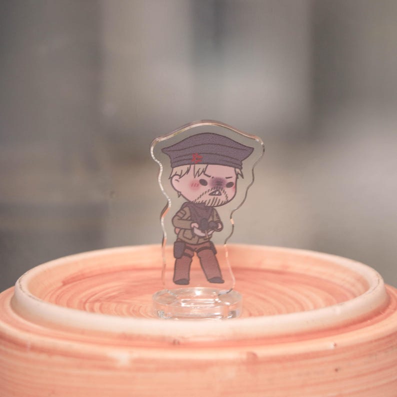May include: A clear acrylic figurine of a cartoon soldier with a stern expression, wearing a dark hat with a red star, a tan uniform, and holding a gun. The figurine is standing on a clear base, set against a blurred background.