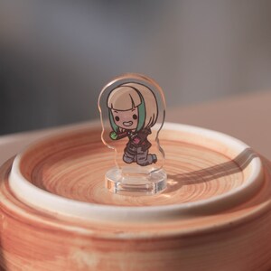 Puede incluir: Una figurita de acr&iacute;lico transparente de un personaje de dibujos animados con cabello rubio, una camisa verde y una chaqueta roja. El personaje sostiene un objeto verde en su mano derecha y est&aacute; de pie sobre una base de acr&iacute;lico transparente.