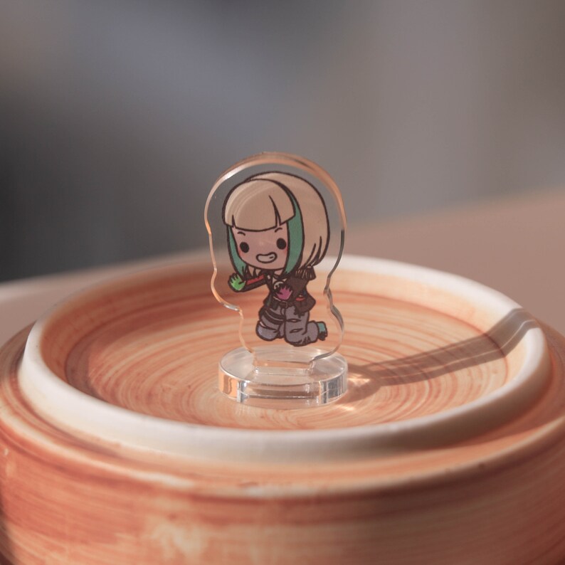 Puede incluir: Una figurita de acr&iacute;lico transparente de un personaje de dibujos animados con cabello rubio, una camisa verde y una chaqueta roja. El personaje sostiene un objeto verde en su mano derecha y est&aacute; de pie sobre una base de acr&iacute;lico transparente.