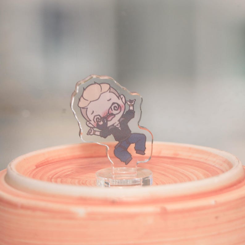 May include: A clear acrylic figurine of a cartoon man with a mustache, wearing a black jacket and blue pants. The figurine is mounted on a clear base and is positioned on a peach-colored, circular object.