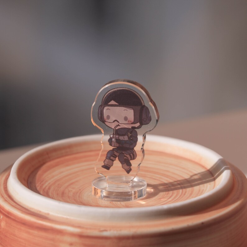 Puede incluir: Una figura de acr&iacute;lico transparente de un personaje de dibujos animados que lleva un atuendo blanco y negro y un casco negro. El personaje sostiene una bebida y est&aacute; sentado sobre una base de cer&aacute;mica marr&oacute;n y blanca.