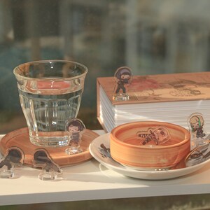 Puede incluir: Una mesa blanca con un vaso de agua, un peque&ntilde;o cuenco de madera, una pila de libros y varias figuritas peque&ntilde;as. Las figuritas son todas personajes diferentes de una caricatura o anime. La mesa est&aacute; puesta sobre una superficie blanca.