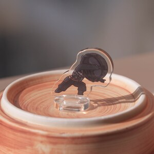 Puede incluir: Una figurita de acr&iacute;lico transparente de un personaje de dibujos animados con un atuendo blanco y negro. La figurita est&aacute; de pie sobre una base de cer&aacute;mica marr&oacute;n y blanca.