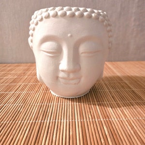 Könnte beinhalten: Weißer Keramik-Pflanztopf in Form eines Buddha-Gesichts mit geschlossenen Augen. Der Topf hat eine glatte, matte Oberfläche und steht auf einer Bambusmatte.