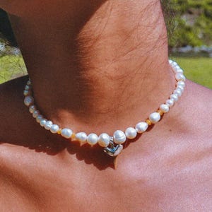 May include: A white pearl necklace with a small silver heart charm. The necklace is on a person's neck.