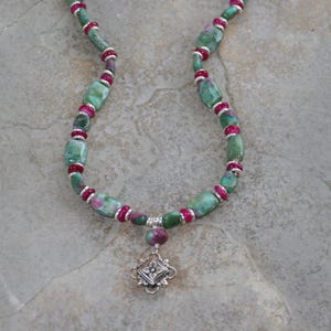 May include: A necklace featuring green and red gemstone beads with silver-colored accents. The necklace has a pendant with a decorative silver-colored charm. The beads are a mix of rectangular and round shapes.