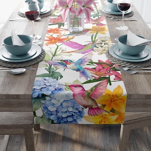 May include: A white table runner with a floral and hummingbird design. The runner features pink, blue, yellow, and green flowers, as well as colorful hummingbirds.