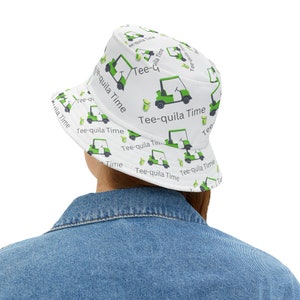 May include: White bucket hat with a repeating pattern of green golf carts and margarita glasses. The text "Tee-quila Time" is printed on the hat. The hat is worn by a person wearing a denim jacket.
