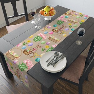 May include: A beige table runner with an Easter theme. The runner features a repeating pattern of pastel colored eggs, green grass, pink tulips, yellow daffodils, and brown bunnies holding baskets of eggs.