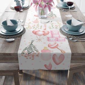 May include: A pink and white baby shower table runner with a pink and white polka dot pattern. The runner features a stork, balloons, a rattle, a baby bottle, and blocks that spell "BABY".
