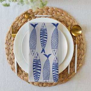 May include: A white plate with a blue and white fish-patterned napkin, gold utensils, and a woven placemat. The napkin features six stylized fish illustrations in blue against a light gray background. The table setting is arranged on a white tablecloth.