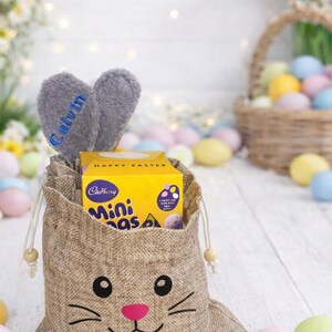 May include: A hessian Easter gift bag with a bunny face design, containing a yellow box of Cadbury Mini Eggs. Grey bunny ears with the name "Calvin" are attached. Pastel Easter eggs and a basket of eggs are in the background.